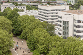 BCI: Rechts im Bild befinden sich zwei hohe graue Gebäude mit vielen Fenstern der Fakultät Bio- und Chemieingenieurwesen. Davor und weiter links im Bild Bäume.